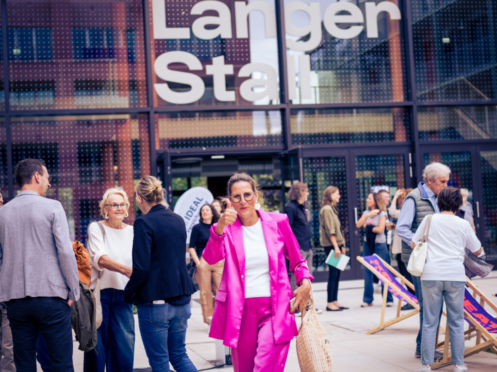 Frau im pinken Anzug zeigt lächelnd den Daumen hoch vor der Fassade „Langer Stall“, im Hintergrund Gäste im Gespräch