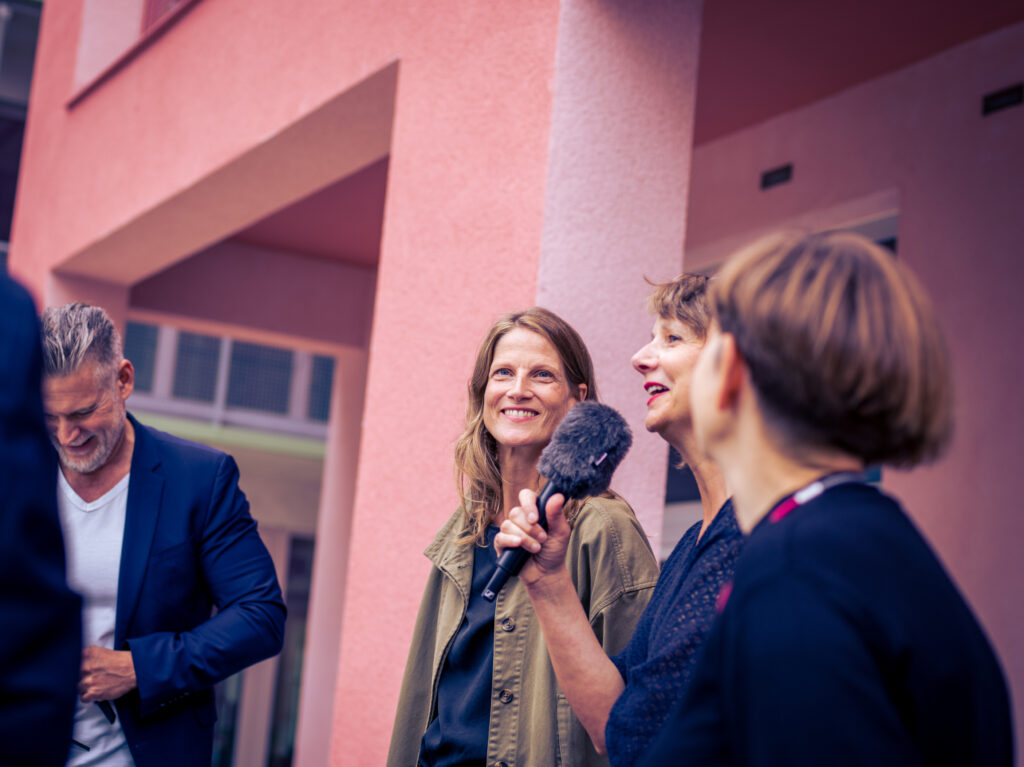 Journalistin interviewt eine lächelnde Frau mit Mikrofon vor der rosafarbenen Fassade