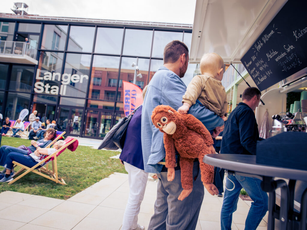 Mann trägt ein Kind und ein Kuscheltier an einem Imbissstand vor der Fassade „Langer Stall“