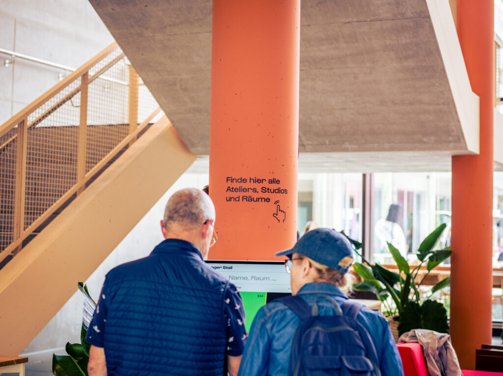 Zwei Besucher informieren sich an einem digitalen Wegweiser-Bildschirm unter der Aufschrift „Finde hier alle Ateliers, Studios und Räume“