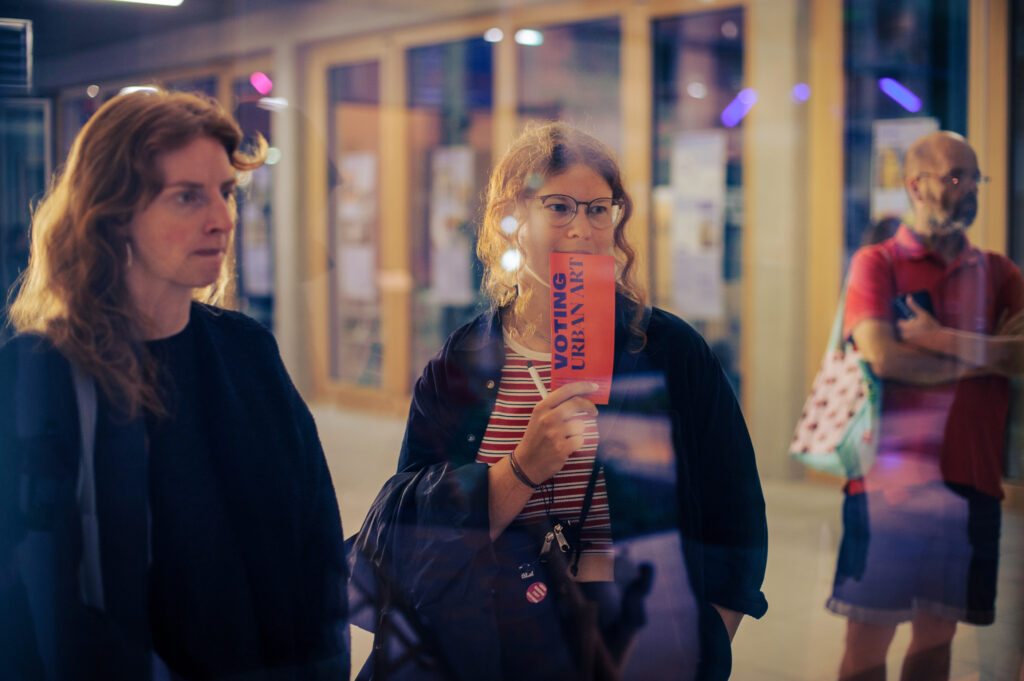 Zwei Frauen betrachten eine rote Abstimmungskarte „Voting Urban Art“ am Abend